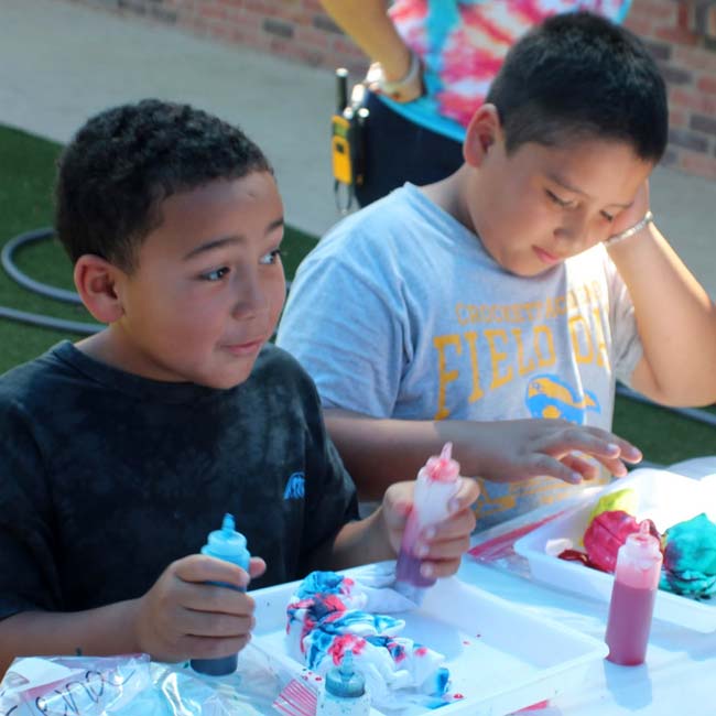 Two boys making tie-dye art outdoors at Lean Valley Daycare, holding bottles and colorful fabric.