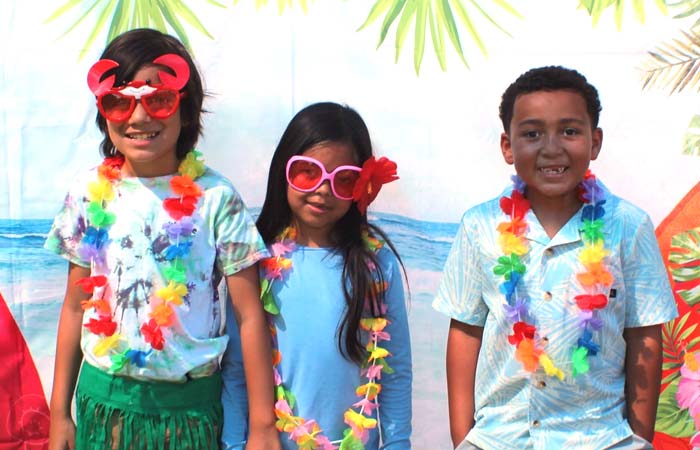 Three preschoolers in leis and sunglasses smile before a tropical beach backdrop.