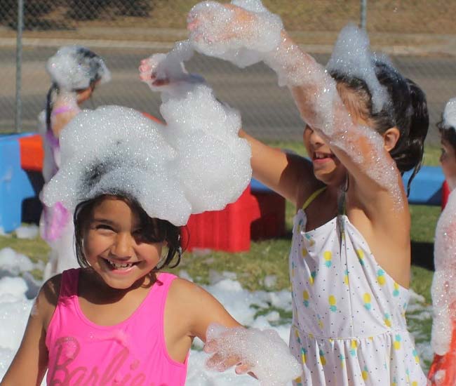 Two girls at Lean Valley Daycare play outside, covered in bubbles and smiling in the sunlight.