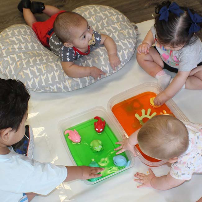 Toddlers at KidCare Learning Center play with colored water and floating toys on a cozy mat.