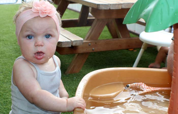 Preschool baby in a tank top stands by a water table with a toy snake, looking surprised.