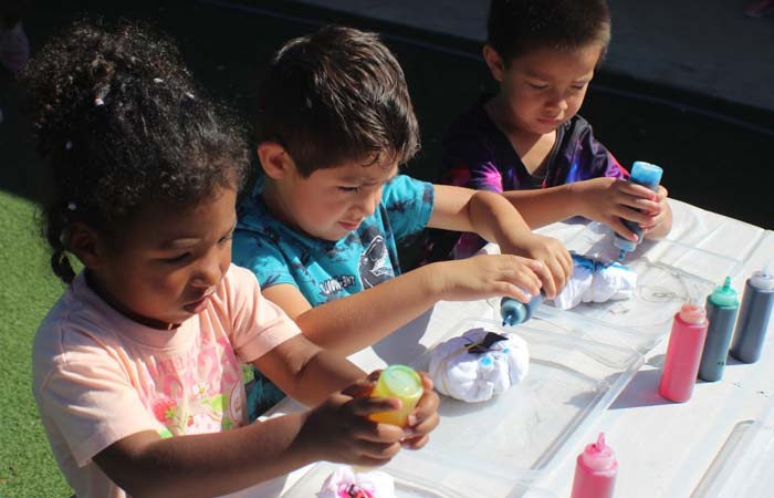 Preschoolers make tie-dye crafts with dye bottles at a sunny outdoor KidCare table.