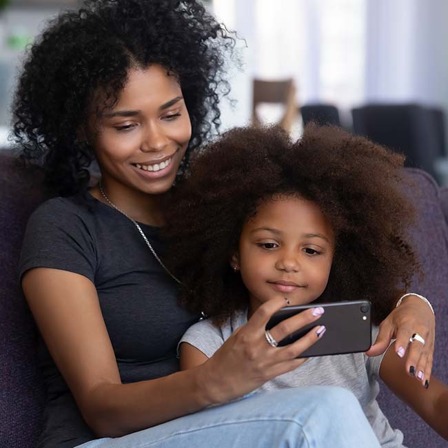 Smiling woman and girl from Lean Valley Daycare sit on couch, taking a selfie with a smartphone.