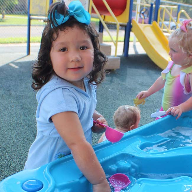 Toddler with blue bow plays at water table near others, enjoying after school playground fun.